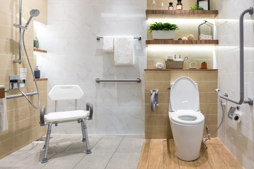 Bathroom designed for accessibility with a shower seat and grab bars for safety. The room features a toilet, shelving, and a tiled floor.