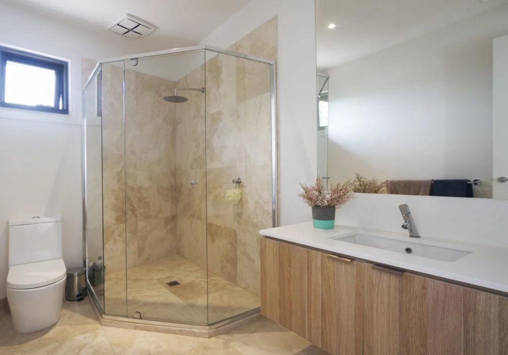 A modern bathroom with a glass shower, toilet, and wooden cabinet with a white sink. The shower features a tan marble-like wall.