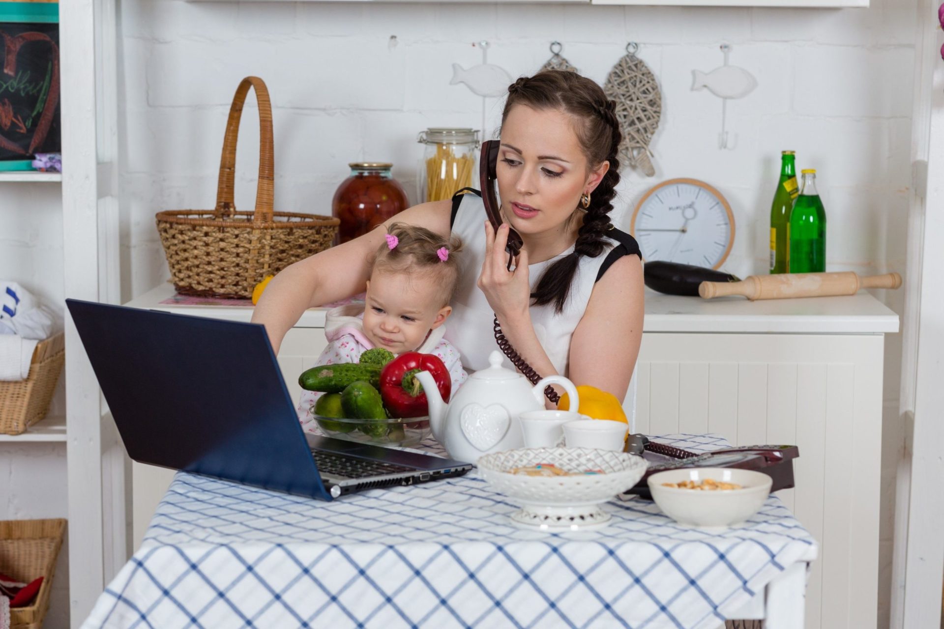 A woman multitasking with a baby on her lap while working on a laptop. The scene is set in a kitchen with a blue and white checkered tablecloth.