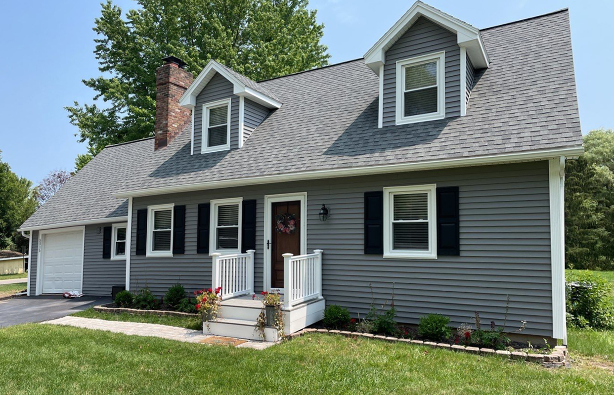 Gray house with white trim and black shutters. The house has a small front porch and a large tree in the background.