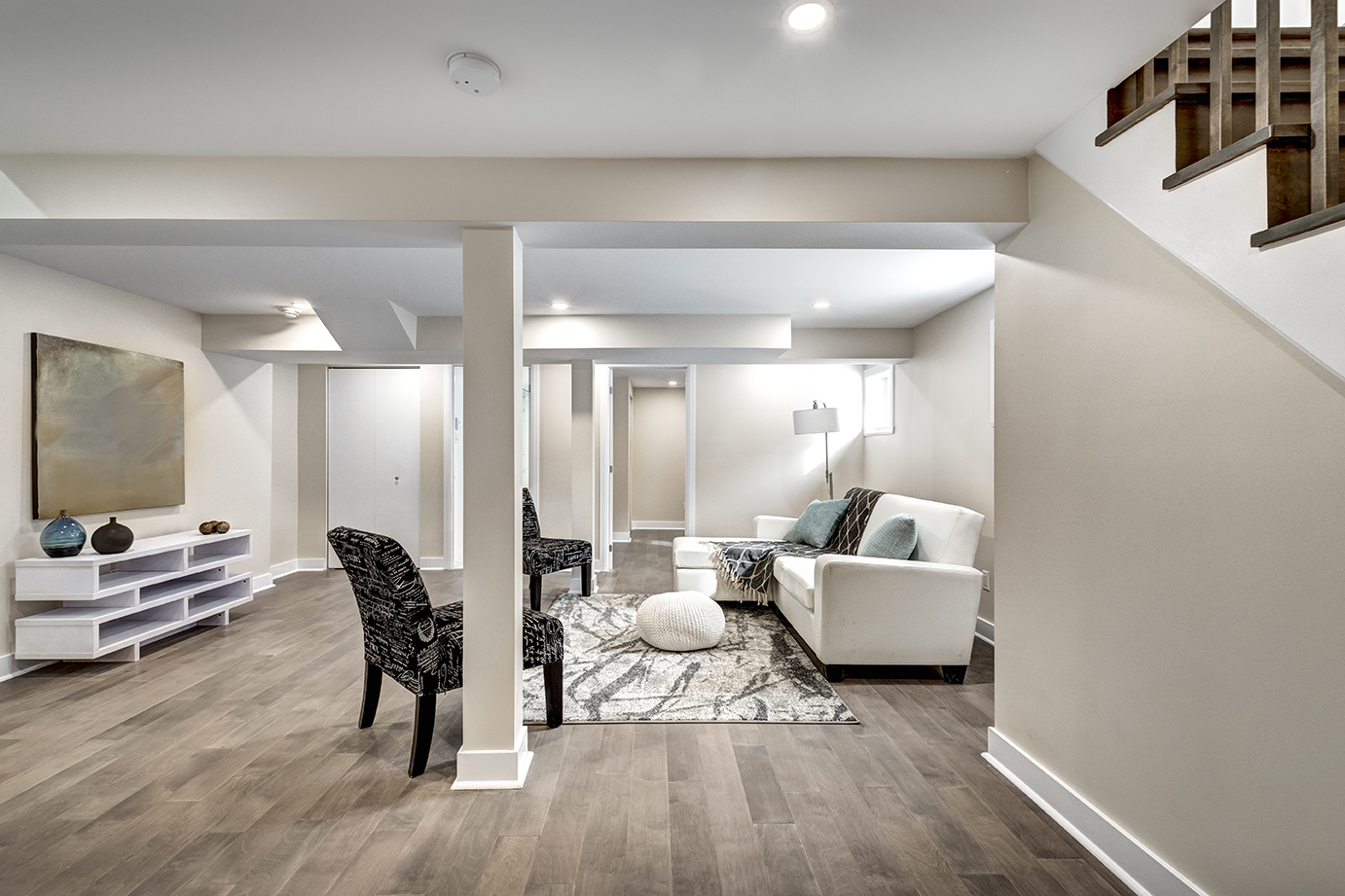 A modern basement living room with white furniture and wooden floors. The room features a white couch and a TV stand.