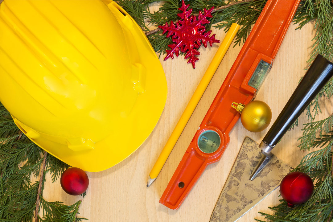 A yellow hard hat, pencil, and orange tool on a table with Christmas ornaments and pine branches.
