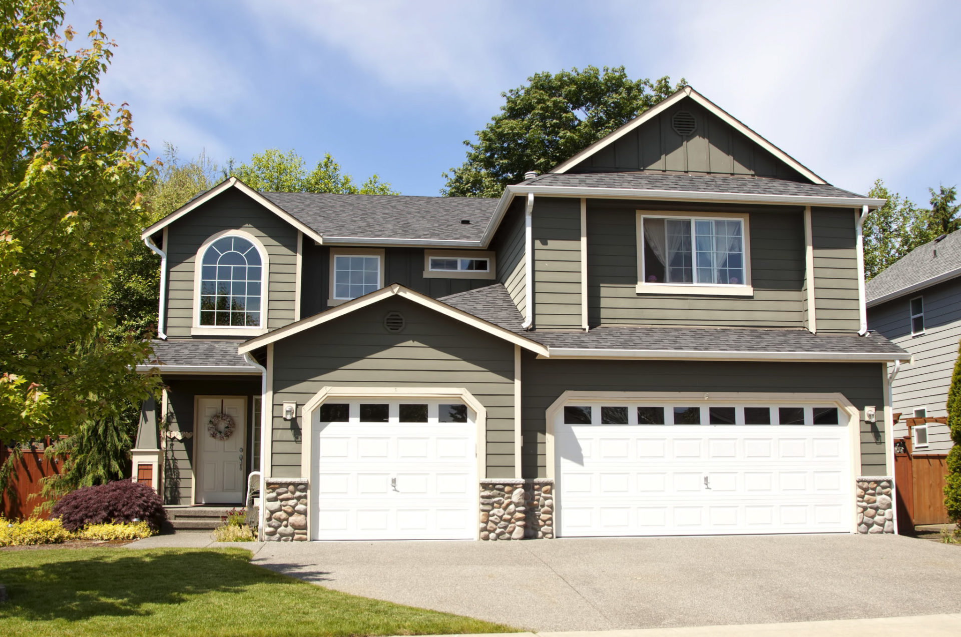 A beautiful two-story house with a large driveway and two white garage doors. The house features gray siding and a stone facade.