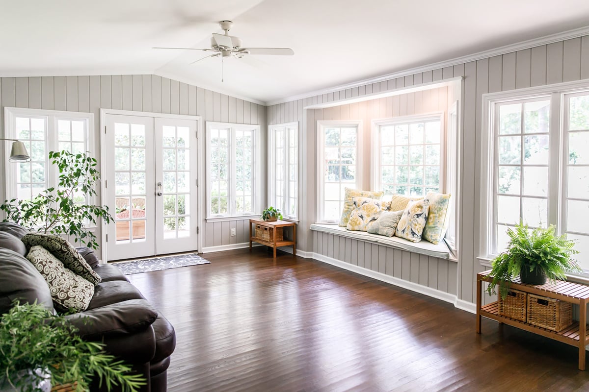 A living room with a brown couch, plants, and a window seat. The room features a dark wood floor and white walls.