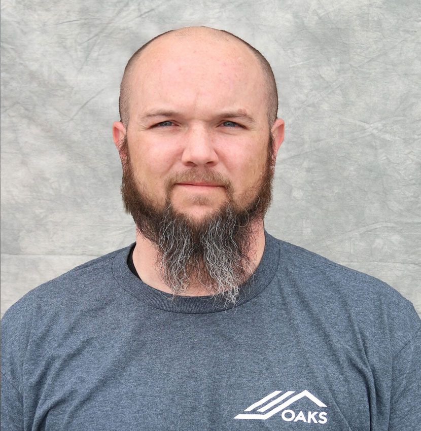 A man with a beard and a OAKS logo shirt. He has a serious expression.