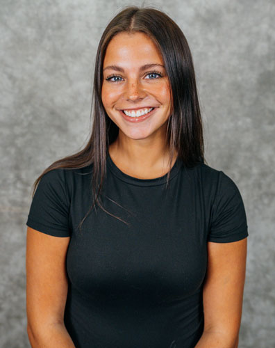 A young woman with long dark hair wearing a black t-shirt smiles at the camera. She has a warm and friendly expression.