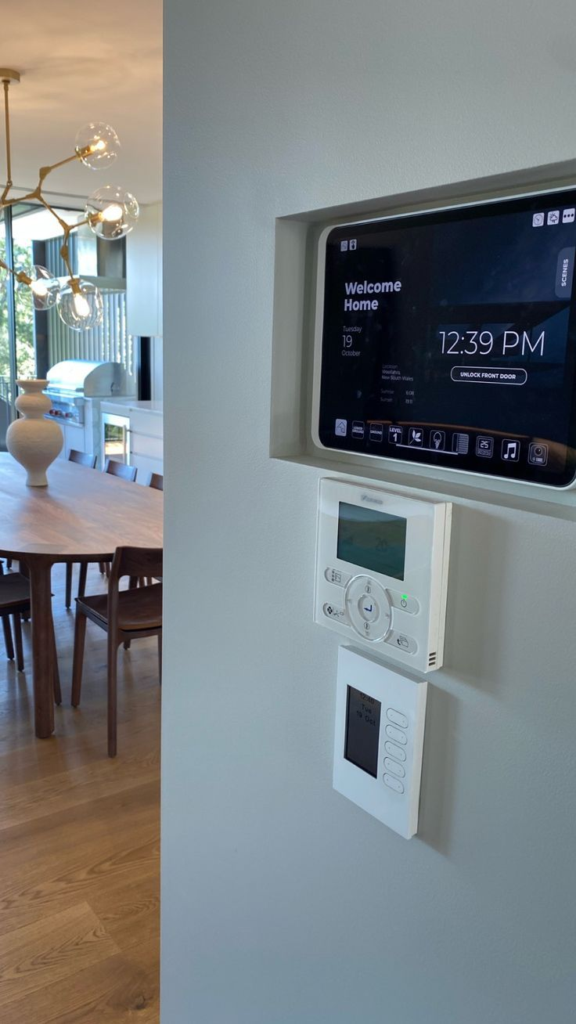 A smart home control panel mounted on a wall with a thermostat below it. The panel displays the time.