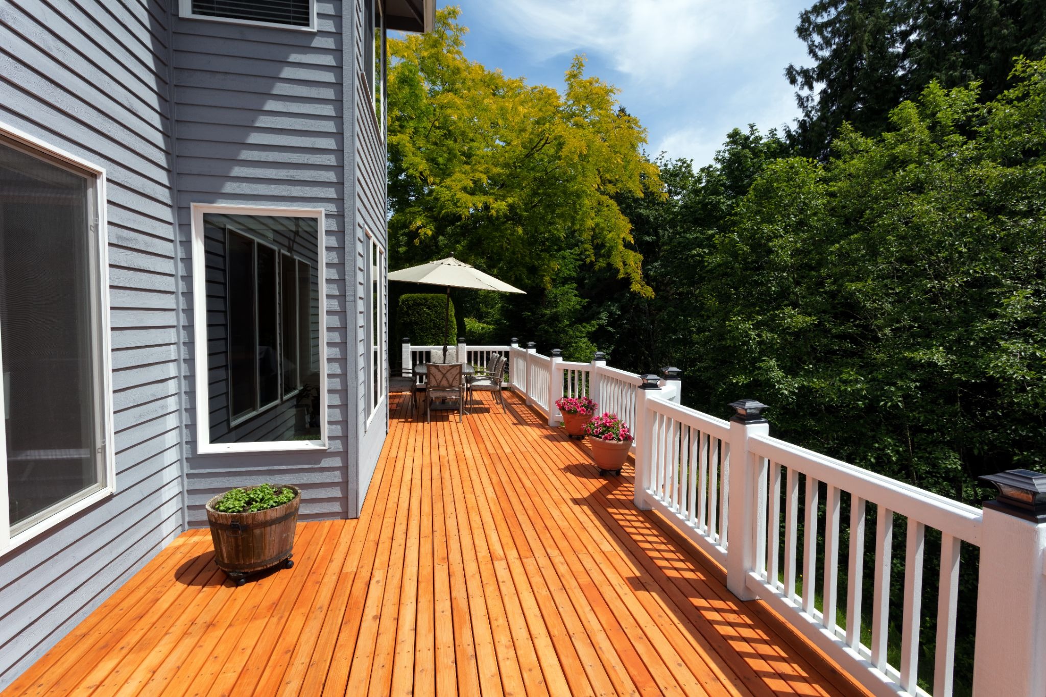 a large wooden deck with white railing and plants. The deck is attached to a house with gray siding.