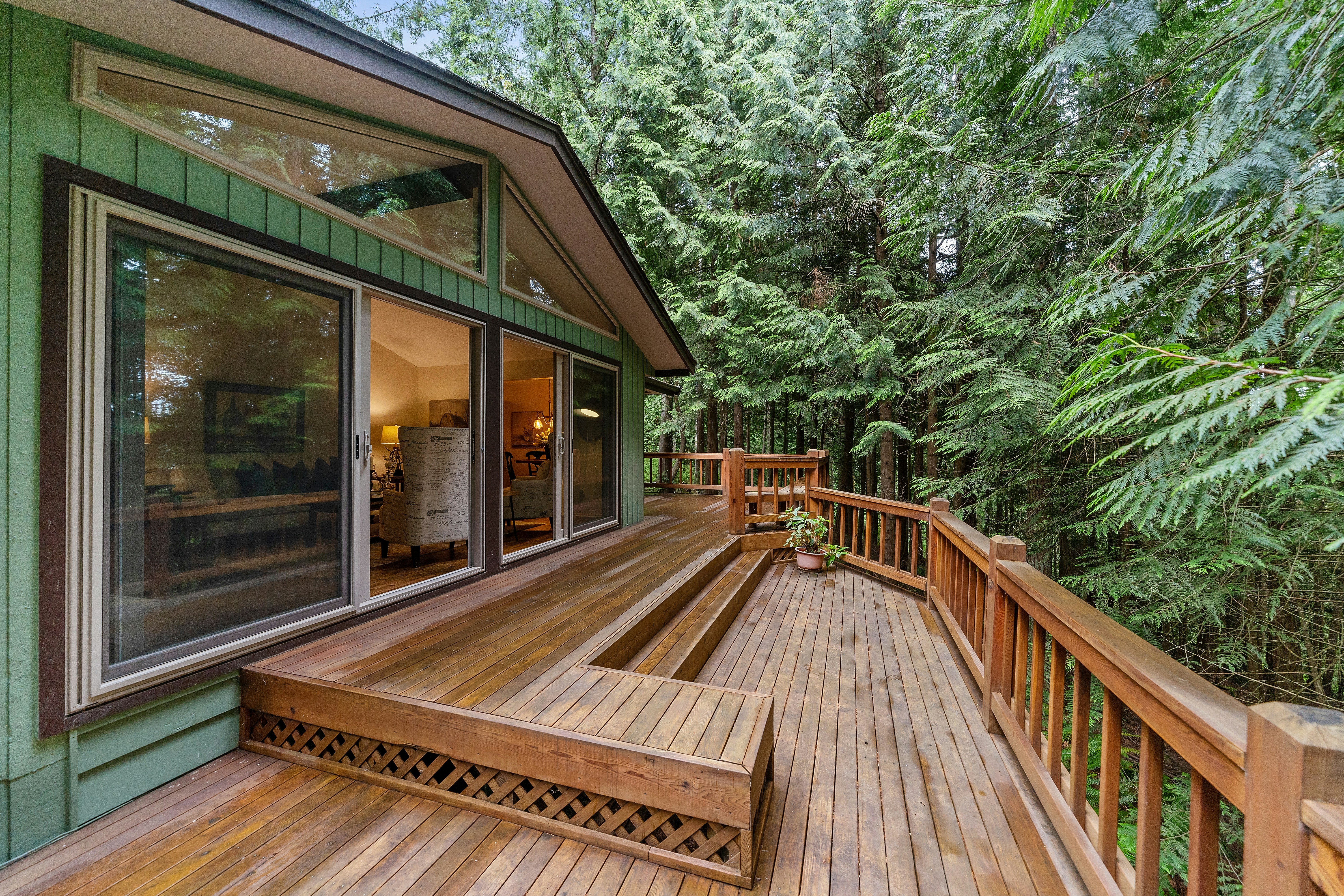 A wooden deck with stairs leading to a green house surrounded by trees. The deck has a railing and a plant on it.