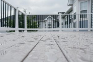 A rain-slicked wooden deck with white railings and a modern house in the background under a cloudy sky.