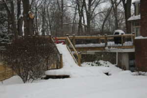 A snowy backyard featuring a wooden deck, ramp, and bare trees in winter.