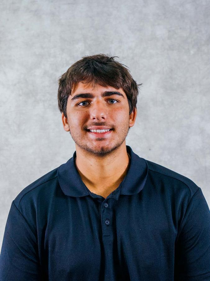 A young man with dark hair and a slight smile, wearing a navy blue polo shirt, is shown in a studio portrait.