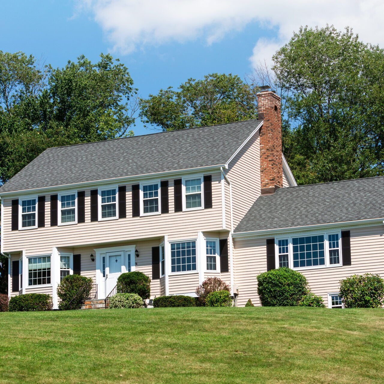A large two-story house with a gray roof and beige siding. The house has a well-manicured lawn and is surrounded by trees.