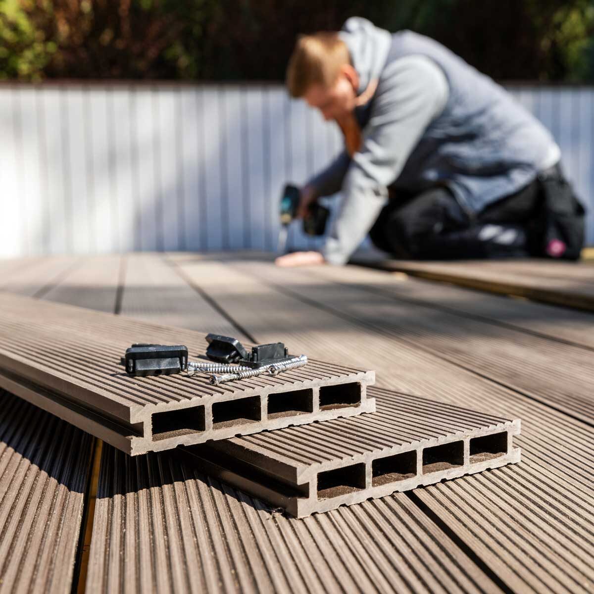 Man kneeling on deck boards, drilling screw, building wooden deck with power tools.