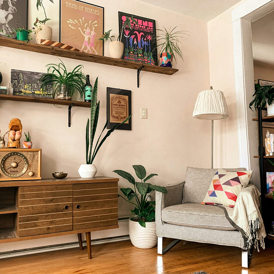 A comfortable living room with various plants and decorative items. The room features a gray chair with a colorful pillow.