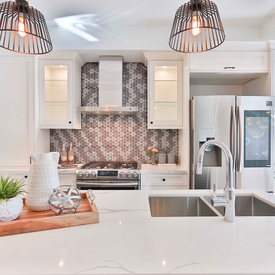 A modern kitchen with white countertops and pendant lights. The kitchen features a stainless steel sink and faucet.