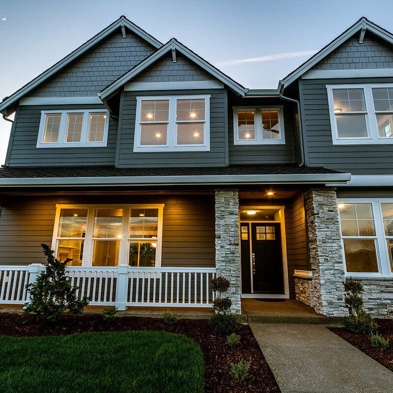A modern two-story house with gray siding and stone accents. The house features a covered porch and a well-manicured lawn.